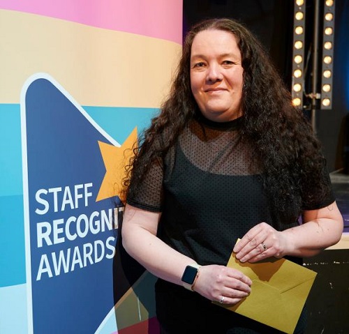 An image of Morag holding her prize envelope and standing beside an upright banner with the award logo to her right 